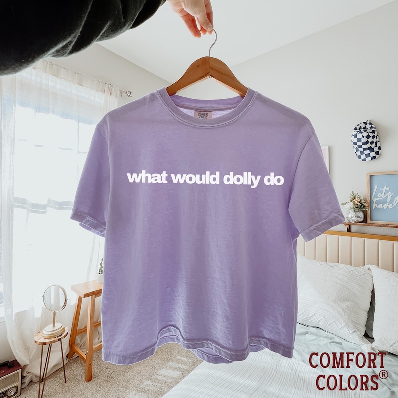 May include: A light purple short-sleeve t-shirt with a crew neckline. The shirt features the white text "what would dolly do" printed across the chest. The garment has a visible Comfort Colors brand tag on the inside of the collar.