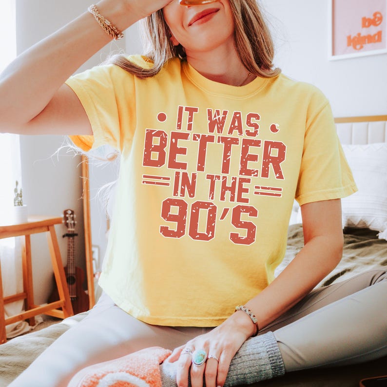 May include: A yellow short-sleeved t-shirt featuring the red distressed text IT WAS BETTER IN THE 90'S printed on the front. The shirt has a crew neckline and a relaxed fit, with the graphic centered on the chest in a bold, block-style font.