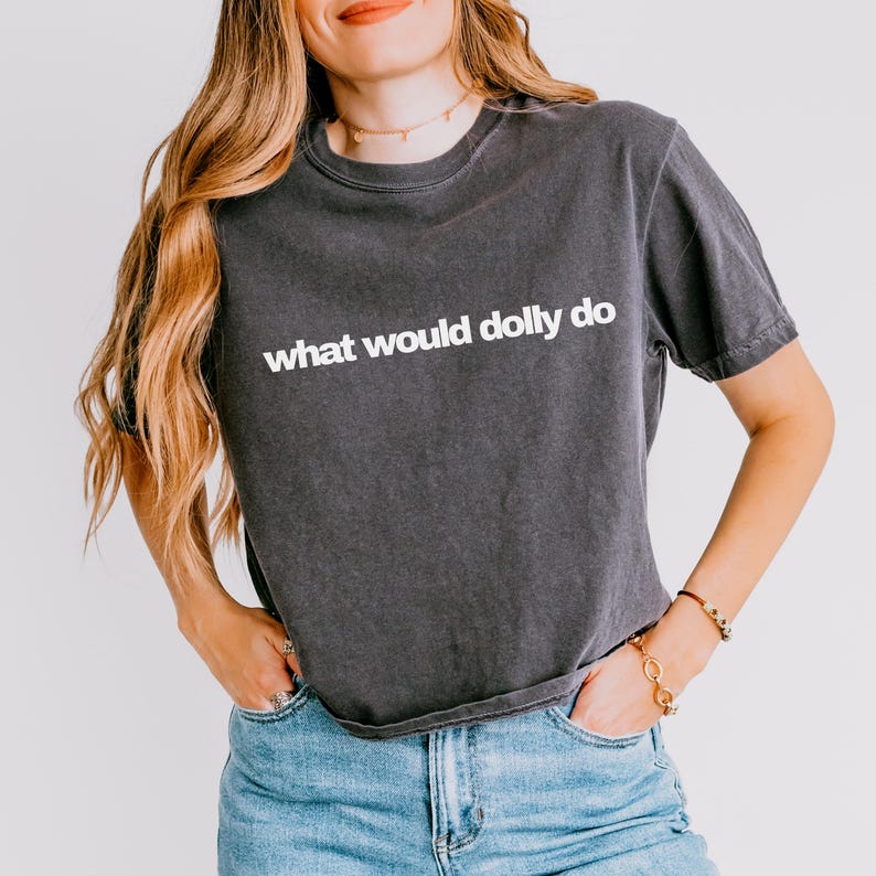 May include: A charcoal gray short-sleeve t-shirt featuring the white text what would dolly do printed across the chest. The shirt has a crew neckline and a relaxed fit, paired with blue denim jeans. The fabric has a slightly faded, vintage appearance.