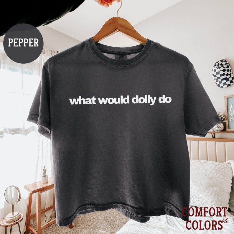 May include: A charcoal gray short-sleeve crew neck t-shirt featuring the white text "what would dolly do" printed across the chest. The shirt is displayed on a wooden hanger and includes a circular tag labeled "PEPPER" in the upper left corner.