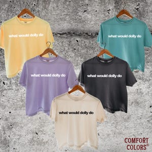 May include: Five short-sleeve crew neck t-shirts in yellow, teal, lavender, charcoal, and cream. Each shirt features the text "what would dolly do" printed on the chest. The shirts are displayed on wooden hangers against a textured gray background.