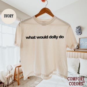 May include: An ivory short-sleeve t-shirt with the black text "what would dolly do" printed across the chest. The shirt features a crew neckline and a standard fit, displayed on a wooden hanger against a bright, neutral-toned indoor setting.
