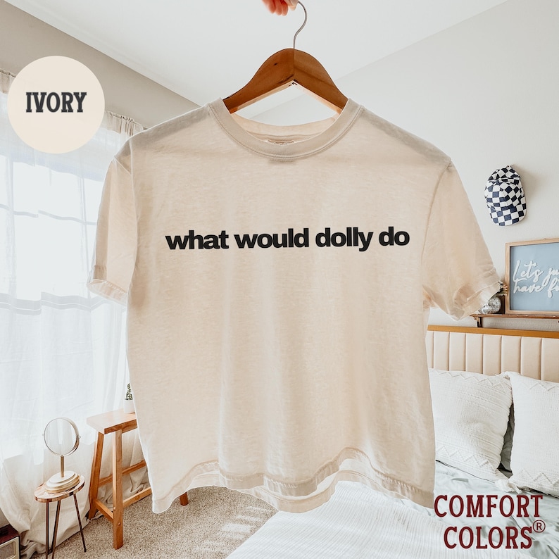 May include: An ivory short-sleeve t-shirt with the black text "what would dolly do" printed across the chest. The shirt features a crew neckline and a standard fit, displayed on a wooden hanger against a bright, neutral-toned indoor setting.