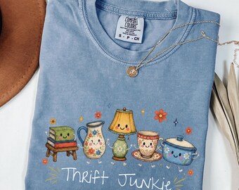 May include: A blue short-sleeve t-shirt with a graphic print of a stack of books, a pitcher, a lamp, a teacup, and a pot, all with smiling faces. The text "Thrift Junkie" is printed below the illustrations in white. The shirt has a Comfort Colors label.