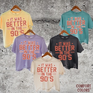 May include: Five short-sleeved crew neck t-shirts in yellow, teal, lavender, charcoal gray, and cream. Each shirt features the red distressed text "IT WAS BETTER IN THE 90'S" printed on the front. The shirts are displayed on wooden hangers.