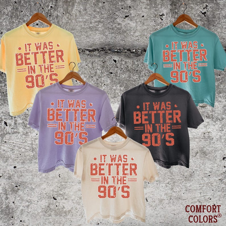 May include: Five short-sleeved crew neck t-shirts in yellow, teal, lavender, charcoal gray, and cream. Each shirt features the red distressed text "IT WAS BETTER IN THE 90'S" printed on the front. The shirts are displayed on wooden hangers.