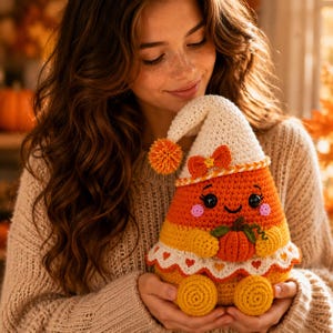 May include: A crocheted candy corn plush toy featuring a white pointed hat with an orange pom-pom, a small orange bow, black button eyes, pink cheeks, and a smiling mouth. The plush holds a small orange pumpkin and has a white scalloped trim.
