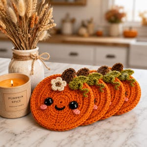 May include: A set of four round orange crochet coasters shaped like pumpkins, each featuring a brown stem, green curly vine, small white flower, black plastic eyes, and pink cheeks. Next to them sits a beige candle labeled Pumpkin Spice.