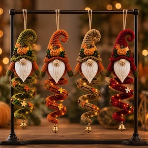 May include: Four hanging crochet gnome ornaments in olive green, orange, tan, and deep red. Each gnome features a pointed hat with a small pumpkin, a white yarn beard, and a long spiral tail decorated with tiny flowers and a small gold bell at the end.