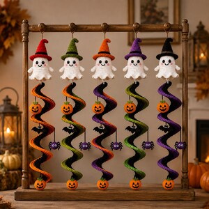 May include: Five crocheted ghost ornaments hanging from a wooden stand. Each ghost wears a different colored witch hat in red, green, orange, purple, or black. Below each ghost is a spiral tail featuring small crocheted pumpkins, bats, and spiders.