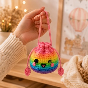 Puede incluir: Una pequeña bolsa redonda de ganchillo con cordón y un degradado de arcoíris. La bolsa tiene una cara guiñando un ojo con ojos de cuentas negras, una boca negra bordada y mejillas rosas. Incluye un asa rosa y dos colgantes en forma de hoja en los extremos de los cordones.