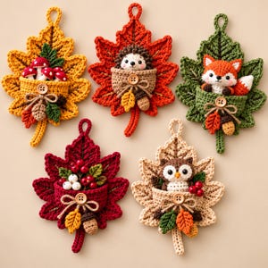 Può includere: Cinque decorazioni da parete all'uncinetto a forma di foglie autunnali nei colori giallo, arancione, verde, rosso e beige. Ogni foglia presenta una piccola tasca con una figura all'uncinetto: funghi, riccio, volpe, bacche o gufo, con bottoni.