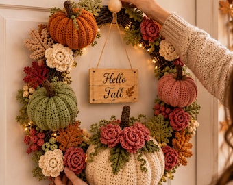 May include: A round wreath featuring crocheted pumpkins in orange, sage green, and cream, accented with crocheted flowers, leaves, and berries. A wooden sign hanging in the center displays the text Hello Fall. The wreath is illuminated by small lights.