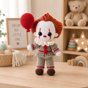 May include: A crocheted doll featuring a white face with black eyes, red facial markings, and orange hair. The doll wears a gray outfit with red pom-poms, a white ruffled collar, and brown shoes, while holding a round, textured red balloon on a string.