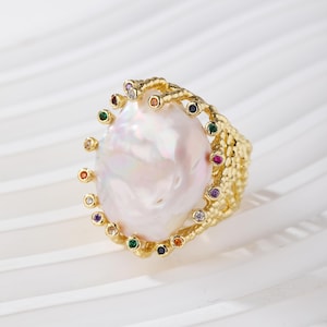 Puede incluir: Un anillo de tono dorado con una gran perla barroca blanca de forma irregular en el centro. La banda tiene una superficie texturizada similar a un panal y está adornada con una serie de pequeñas piedras preciosas multicolores alrededor.