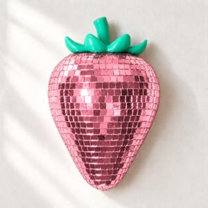 May include: A strawberry-shaped disco ball covered in small, reflective pink mirror tiles. The top of the strawberry features a smooth, bright green stem and leaf structure, creating a vibrant contrast with the shimmering pink body.