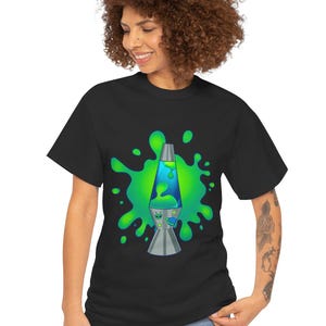May include: A black short-sleeve t-shirt featuring a graphic print of a lava lamp centered against a bright green liquid splash. The lamp contains blue liquid and has a small alien face and a flying saucer icon on its silver base.