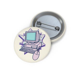 May include: A round pin-back button featuring a light-colored illustration of a vintage desktop computer, keyboard, and mouse. The computer screen is light blue, set against a purple starburst graphic with small geometric shapes on a cream background.