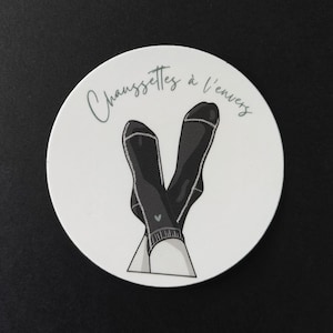 May include: A circular white sticker featuring a graphic illustration of two crossed dark gray socks with a small heart detail. The text "Chaussettes à l'envers" is printed in a dark gray cursive script along the top curve of the circular sticker.
