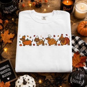 Könnte beinhalten: Ein weißes Sweatshirt mit Rundhalsausschnitt und einer horizontalen Grafik von vier Dackeln in herbstlichen Kostümen, darunter ein Kürbis, ein Kuchen, ein gebratenes Truthahn-Gericht und ein Truthahn-Kostüm, umgeben von Blättern und Eicheln.