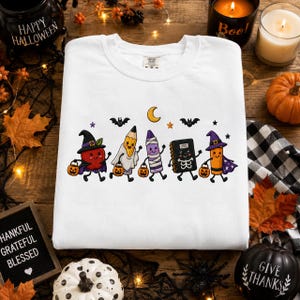 Könnte beinhalten: Ein weißes Sweatshirt mit Rundhalsausschnitt und einer bunten Grafik von fünf anthropomorphen Schulsachen in Halloween-Kostümen, darunter eine Hexe, ein Geist, eine Mumie, ein Skelett und ein Zauberer, die kleine orangefarbene Kürbiseimer tragen.
