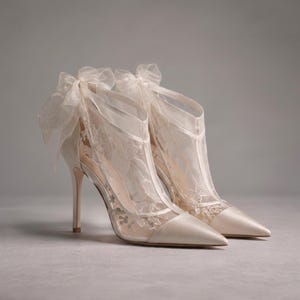 Könnte beinhalten: Ein Paar elfenbeinfarbene High-Heel-Pumps mit spitzer Zehenpartie, Satin-Finish und einem transparenten Netzüberzug mit zarter Blumenstickerei. Die Schuhe sind mit einer großen, transparenten Organzaschleife an der Ferse und einem schmalen Stiletto-Absatz versehen.