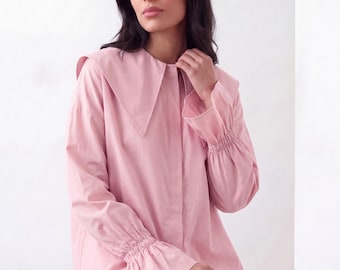 May include: A light pink long-sleeve button-down shirt featuring a large pointed collar and gathered cuffs with flared sleeve ends. The shirt is paired with white trousers, creating a clean and simple look with soft pastel tones.
