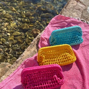 May include: Three rectangular crochet pouches in bright turquoise, yellow, and hot pink. Each pouch features an open-weave net pattern and a structured frame closure. The pouches are arranged in a diagonal line on a textured pink fabric surface.