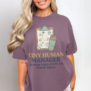 Può includere: Una t-shirt a maniche corte color malva con la grafica di un blocco per appunti con omini stilizzati e una tazza da viaggio. Il testo recita: TINY HUMAN MANAGER. Meetings begin at 8:00 AM. Nobody listens. Toni tenui di verde, blu e giallo.