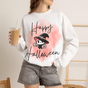 Könnte beinhalten: Ein weißes Langarm-Sweatshirt mit einem rosa Aquarell-Spritzer-Motiv und dem schwarzen Text Happy Halloween. Das Design zeigt eine Cartoon-Figur mit einem schwarzen Hexenhut, die einen kleinen Totenkopf und einen Kürbis hält.