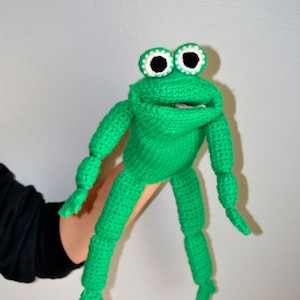 Peut inclure: Marionnette de grenouille en crochet vert avec des yeux blancs et une grande bouche ouverte. La marionnette est tenue par une main.