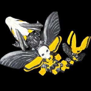 May include: A 3D printed articulated insect figurine featuring a white skull-shaped head and thorax, large black wings with yellow accents, and a segmented body colored in alternating black and yellow segments, displayed against a black background.