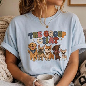 May include: A light blue short-sleeve t-shirt featuring a graphic print of five dogs and the text "THE GROUP CHAT" in colorful, retro-style lettering. The dogs depicted include a golden retriever, a French bulldog, a corgi, a dachshund, and a chihuahua.