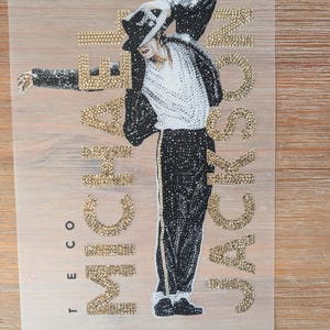 Pode incluir: Um design de transferência de strass com uma figura de chapéu, casaco e calças pretas com uma camisa branca, em pose de dança. O texto MICHAEL JACKSON é formado por strass dourados, com o texto vertical TE CO em pequenos strass dourados.