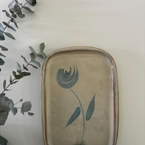 May include: A rectangular ceramic tray with rounded corners, featuring a beige glazed surface and a hand-painted blue flower design in the centre. The edges of the tray have a warm brown tone, and the overall finish appears smooth and glossy.