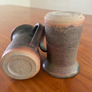 May include: Two cylindrical ceramic mugs with a dark, textured, mottled brown and charcoal finish. One mug is positioned upright, while the other lies on its side, revealing a circular base with an impressed maker's mark and a handle.
