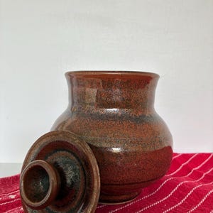 May include: A round, bulbous ceramic jar with a glossy, mottled finish in shades of deep rust, brown, and black. The jar features a wide opening and a matching circular lid with a central knob and a small notch for a spoon or utensil.