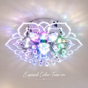 以下が含まれることがあります： 蓮の花の形をした天井照明器具で、透明なアクリルの花びらと多数の吊り下げられたクリスタル球が特徴です。光はマルチカラーの輝きを放ち、画像の下部には「Exposed Color-Turn on」というテキストが印刷されています。