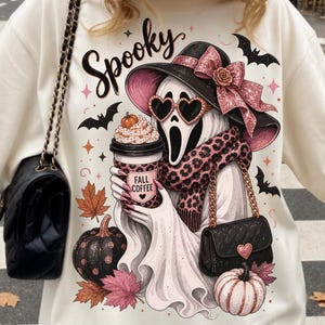 May include: A cream-colored t-shirt featuring a graphic of a ghost wearing a black hat with a pink glittery bow, heart-shaped sunglasses, and a leopard print scarf. The ghost holds a coffee cup labeled Fall Coffee, surrounded by bats and pumpkins.