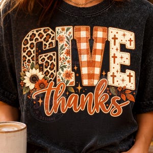 Könnte beinhalten: Ein schwarzes T-Shirt in Acid-Wash-Optik mit dem Schriftzug GIVE THANKS in großen Blockbuchstaben. Die Buchstaben sind mit Leopardenmuster, Blumenmustern, orangefarbenem Vichy-Karo und Kreuzmotiven verziert. Das Wort thanks ist in orangefarbener Schreibschrift gehalten.