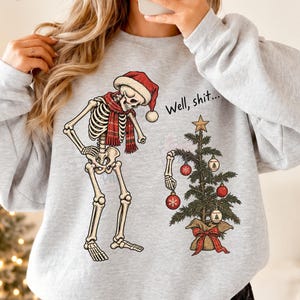 Könnte beinhalten: Ein hellgraues Sweatshirt mit Rundhalsausschnitt und einer Grafik eines Skeletts mit roter Weihnachtsmütze und Schal, das einen kleinen geschmückten Weihnachtsbaum betrachtet. Der Text "Well, shit..." steht über dem Baum mit roten und weißen Kugeln.