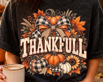 May include: A charcoal grey mineral-wash t-shirt featuring a graphic print of pumpkins, sunflowers, autumn leaves, and a black and white buffalo plaid bow. The text THANKFULL is printed in large, bold, off-white letters across the center of the design.