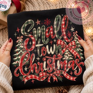 May include: A black sweatshirt featuring the text Small Town Christmas in red and green plaid script lettering. The design includes illustrations of two Christmas trees, red bows, and small stars, creating a festive holiday graphic.