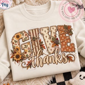 Könnte beinhalten: Ein cremefarbenes Sweatshirt mit Rundhalsausschnitt und dem Schriftzug GIVE THANKS im Patchwork-Stil. Die Buchstaben sind mit Sonnenblumenmustern, braunen Streifen, Kreuzsymbolen und Herbstblättern verziert sowie mit Garn und Knöpfen akzentuiert.