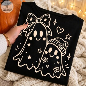 Könnte beinhalten: Ein schwarzes T-Shirt mit einer hellen Grafik von zwei Geistern. Ein Geist trägt eine große Schleife, der andere eine Mütze mit einem Smiley-Gesicht. Das Design enthält kleine Sterne, Herzen und Blumenformen, die die Geister umgeben.
