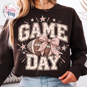 Könnte beinhalten: Ein dunkelbraunes Langarm-Sweatshirt mit einem Grafikdruck, der den Text GAME DAY in großen Blockbuchstaben mit hellrosa Punkten zeigt. Ein brauner Football mit einer Seite im Leopardenmuster und einer großen hellrosa Schleife ist mittig.