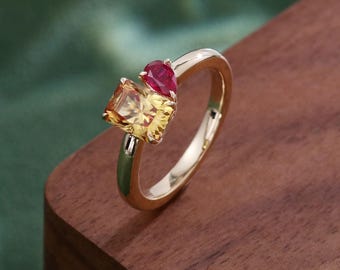 May include: A gold ring featuring a rectangular yellow gemstone and a smaller pear-shaped red gemstone set side-by-side. The band is polished gold, showcasing a smooth, reflective surface that complements the vibrant colors of the two faceted stones.