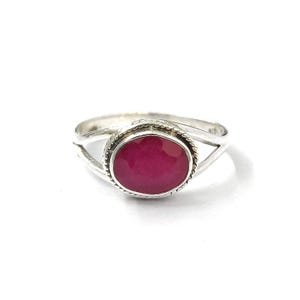 May include: A silver ring featuring a central oval-shaped, faceted deep pink gemstone. The stone is set within a twisted rope-style metal border, and the band splits into two separate silver strands on each side of the setting, creating an open design.