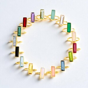May include: A collection of gold-toned rings arranged in a circle, each featuring a single rectangular baguette-cut gemstone in various colors including red, purple, green, blue, yellow, pink, black, and clear, set on a thin metallic band.