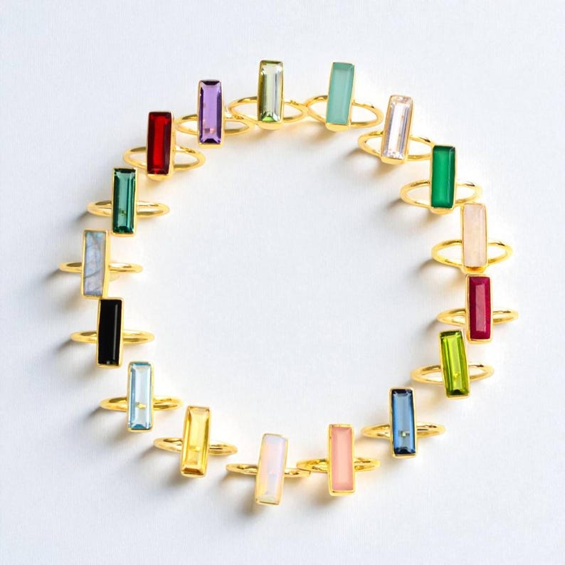 May include: A collection of gold-toned rings arranged in a circle, each featuring a single rectangular baguette-cut gemstone in various colors including red, purple, green, blue, yellow, pink, black, and clear, set on a thin metallic band.